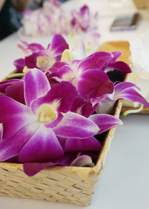 Close-up of a purple orchid blossom resting inside a woven lauhala basket placed on a table, highlighting the flower’s vibrant color and the basket’s textured weave.