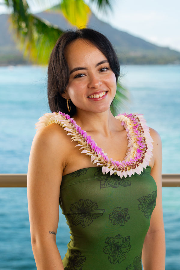 A young woman in her 20s wearing a dark green dress with black floral patterns and a pastel pink flat orchid lei, standing in front of Diamond Head, a palm tree, and the ocean