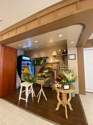 A quaint corner lei shop featuring a table with floral arrangements, a fridge filled with leis, a person opening the fridge to select a lei, a stool, a glass display case, and fresh leis hanging in the background.