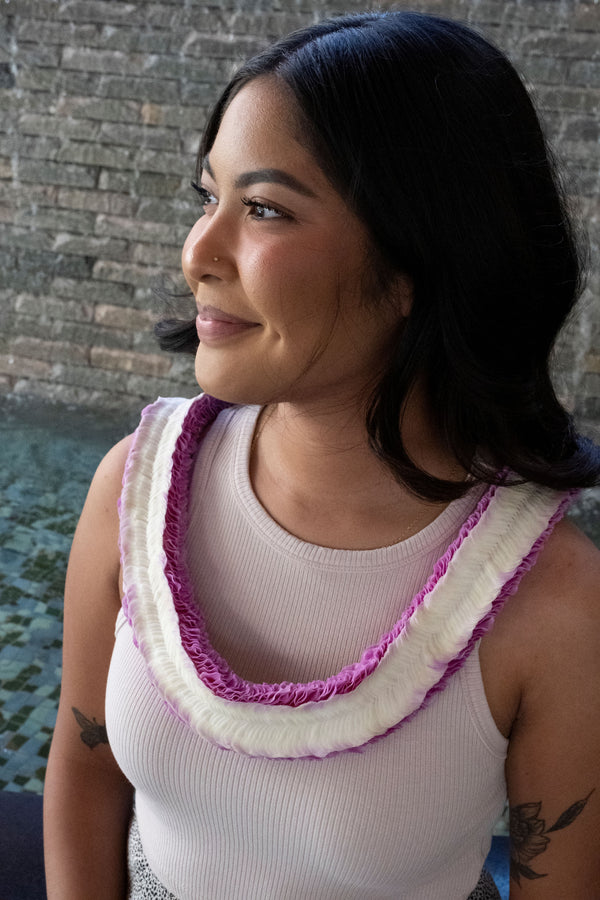 A woman in a beige tank top wearing a white and pink orchid lei, standing in front of a grey brick background.