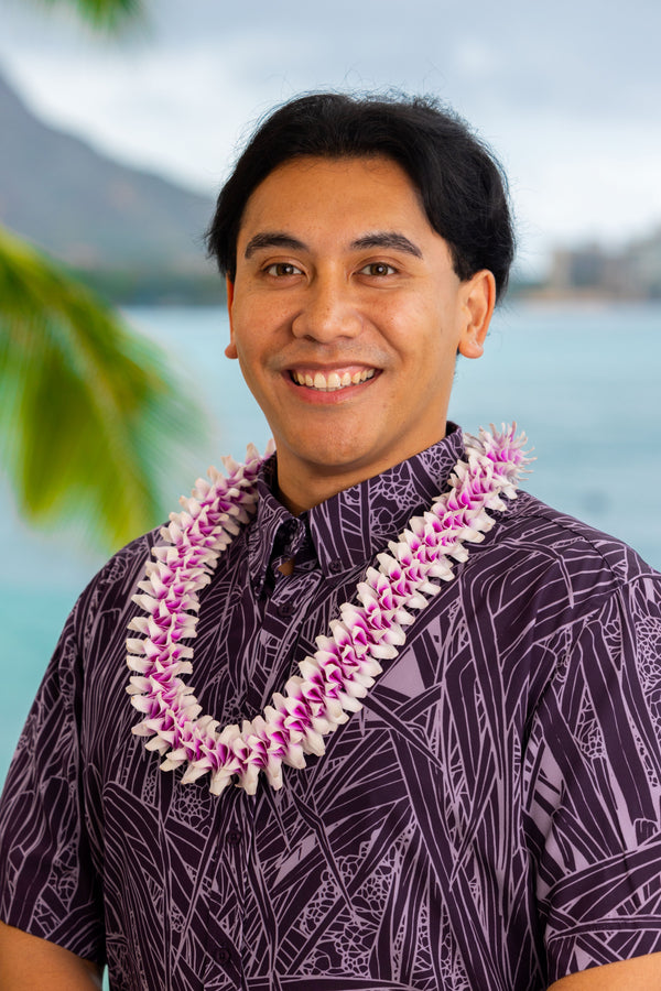 Man wearing a purple patterned shirt and a pink and purple orchid spiral lei, with Diamond Head, a palm tree, and the ocean in the background.