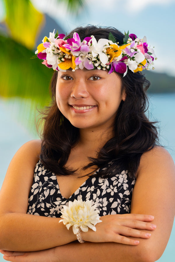 Girl wearing a multicolored orchid and green fern lei po‘o, with greenery and ocean in the background.