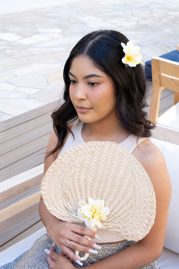Girl wearing a silk yellow plumeria hair clip, holding a woven fan decorated with silk plumeria flowers, sitting on a bench.