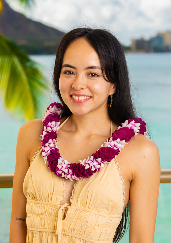 A woman wearing a light yellow dress and a two-shaded purple spiral orchid lei, standing with Diamond Head, a palm tree, and the ocean in the background.