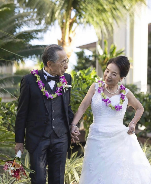 Elderly wedding couple wearing single-strand orchid leis in purple and green, standing together with a tropical background of palm trees and lush greenery.