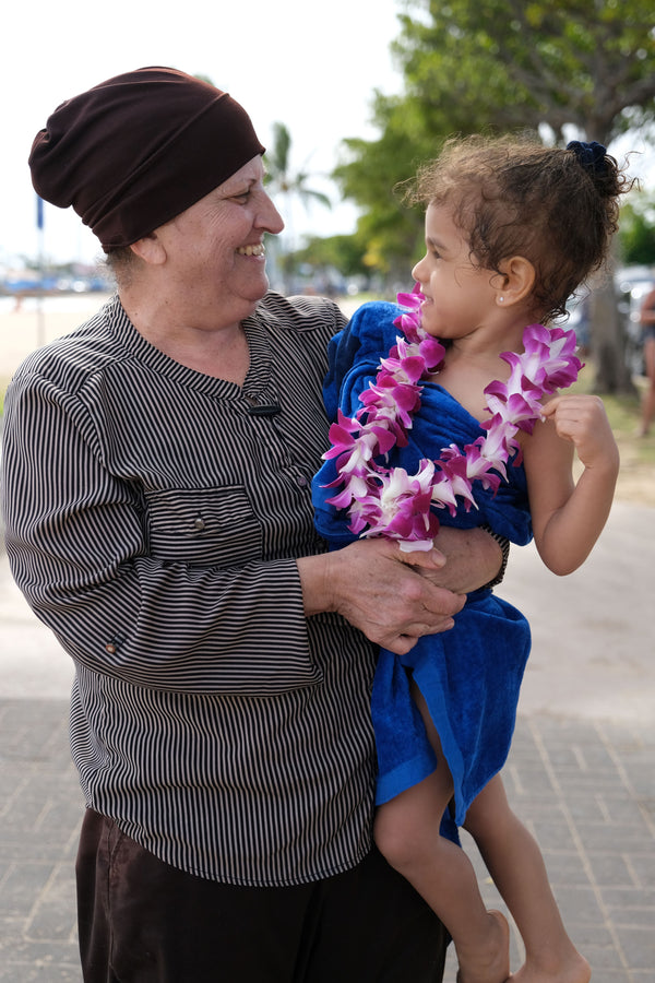 Elderly woman holding a young girl wearing a kid-size single strand purple orchid lei, with the beach in the background.