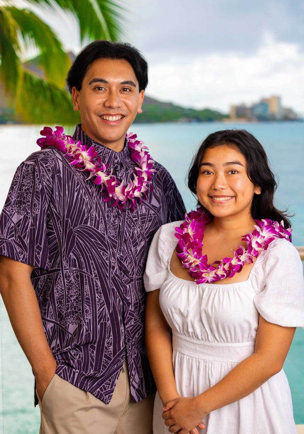 A man and woman standing side by side, each wearing a classic single purple orchid lei, with Diamond Head, a palm tree, and the ocean in the background.