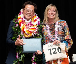 The owner of the orchid lei company on the left wearing multiple leis, holding up his pineapple award, standing next to a woman on the right who is also wearing a lei and holding up a Hawaii Gas pouch.