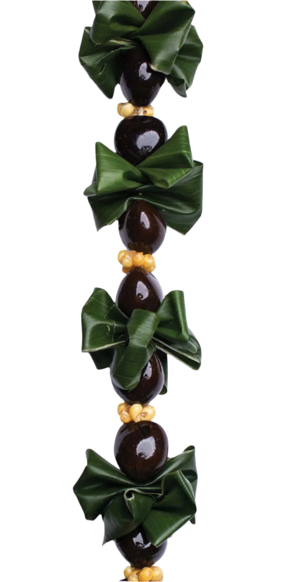 Close-up of a ti leaf kukui nut lei with folded green ti leaves, shiny black kukui nuts, and white mongo shells, highlighting the texture and color contrast against a white background.