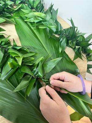 Person crafting a green ti leaf maile lei using fresh ti leaves on a wooden table