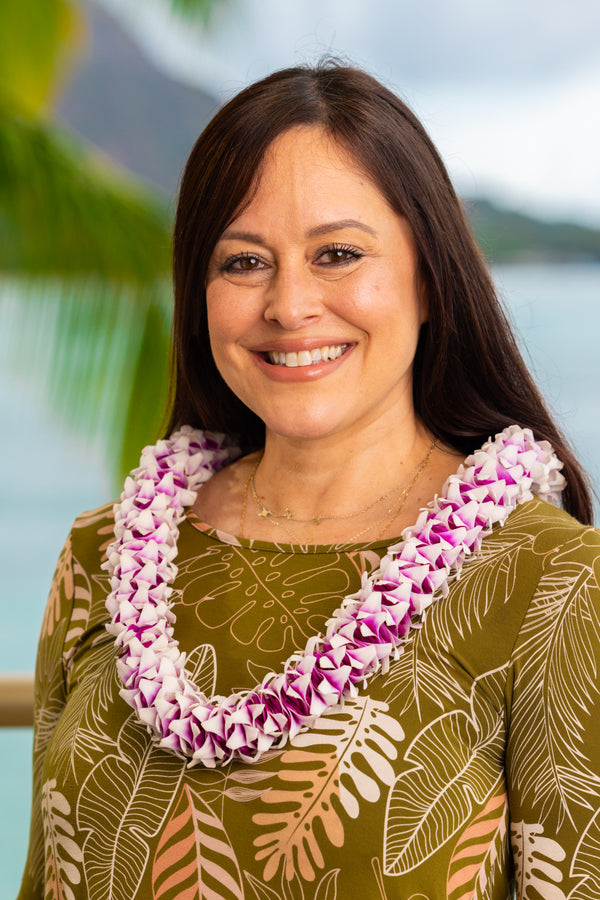 Woman wearing a green leaf design top and a textured purple spiral orchid lei, with Diamond Head, a palm tree, and the ocean in the background.
