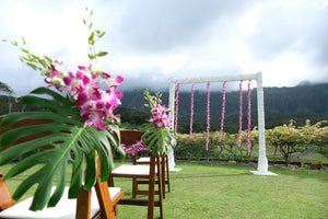A lush green outdoor Hawaiian venue featuring an orchid garland-decorated arch and wooden chairs adorned with monstera leaves and orchid stems.