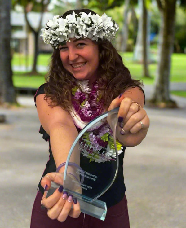 Person holding an award outdoors, wearing a white orchid and green fern lei po‘o, with greenery in the background.