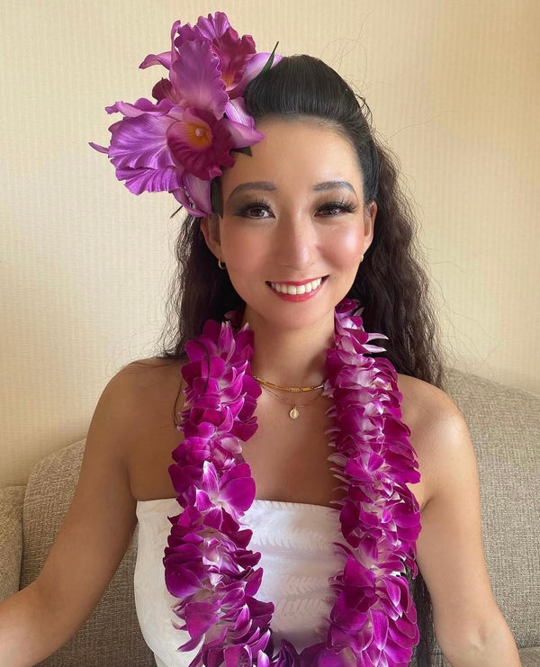 Woman sitting on a couch wearing a purple floral headpiece and a fluffy purple orchid lei.