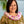 A woman wearing a green leafy pattern shirt and a fluffy deluxe orchid lei made of pink, purple, and white blossoms, standing in front of Diamond Head with a palm tree and the ocean in the background.