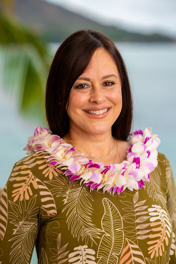A woman wearing a green leafy pattern shirt and a fluffy deluxe orchid lei made of pink, purple, and white blossoms, standing in front of Diamond Head with a palm tree and the ocean in the background.