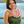 A young woman in her 20s wearing a dark green dress with black floral patterns and a pastel pink flat orchid lei, standing in front of Diamond Head, a palm tree, and the ocean