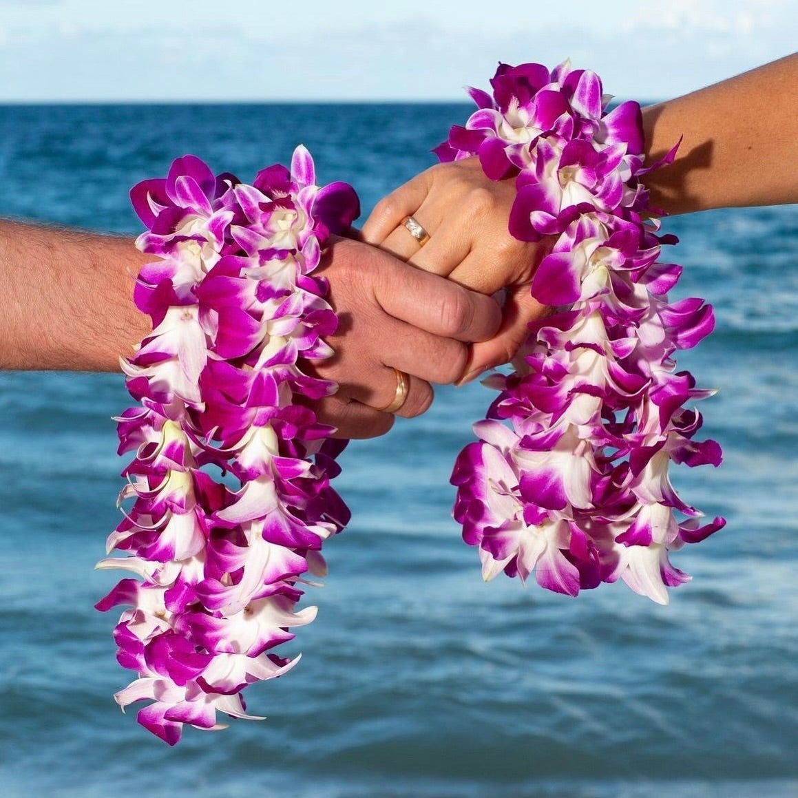 A married couple holding hands, each wearing a single-strand purple orchid lei draped around their hands.