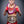 A woman in boxing attire holding a championship belt, wearing a white and purple layered orchid lei. She stands confidently against a neutral gray background, highlighting the vibrant lei and her athletic achievement.