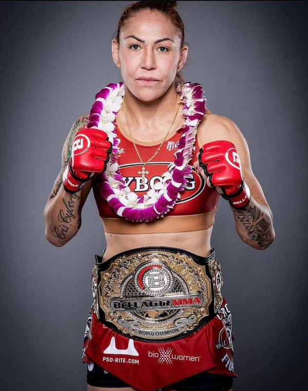 A woman in boxing attire holding a championship belt, wearing a white and purple layered orchid lei. She stands confidently against a neutral gray background, highlighting the vibrant lei and her athletic achievement.