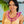 A young woman in a light yellow dress wearing a flat deluxe lei made of purple orchids, with Diamond Head, a palm tree, and the ocean in the background.