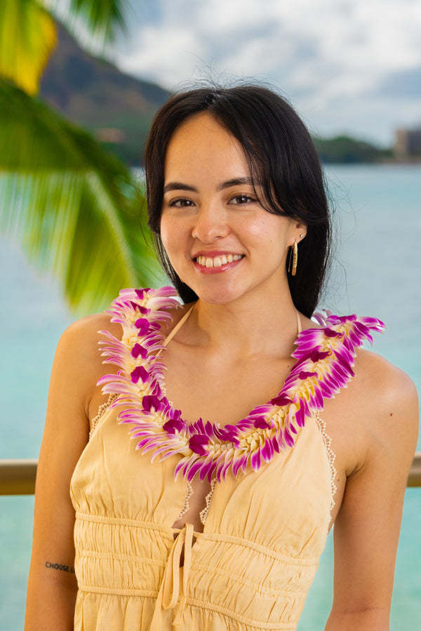 A young woman in a light yellow dress wearing a flat deluxe lei made of purple orchids, with Diamond Head, a palm tree, and the ocean in the background.