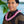 A man wearing a black and brown patterned shirt with a textured purple orchid lei featuring a loop-like folding design, with Diamond Head, a palm tree, and the ocean in the background.
