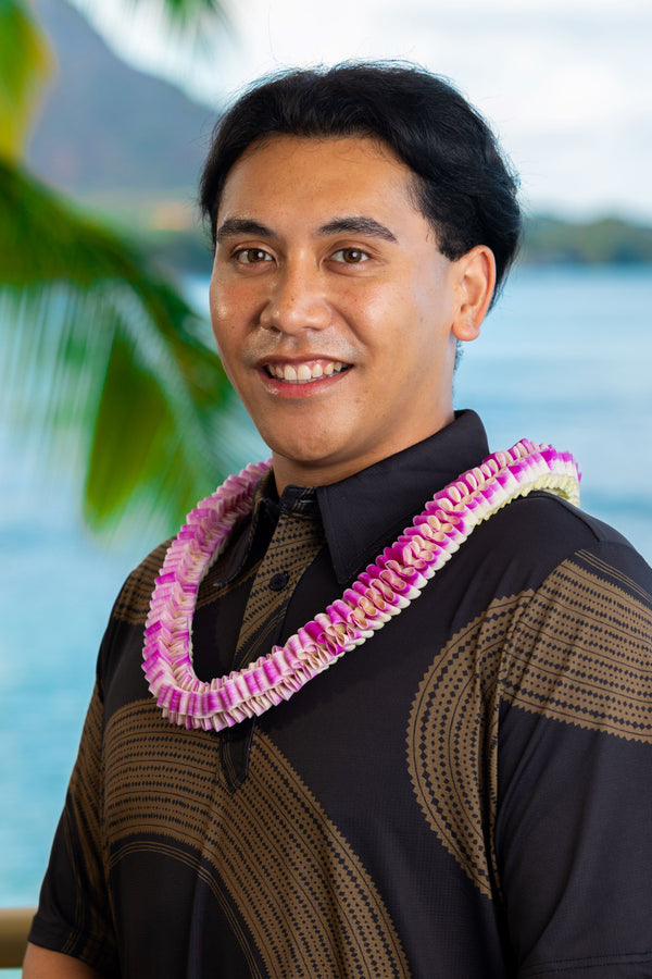 A man wearing a black and brown patterned shirt with a textured purple orchid lei featuring a loop-like folding design, with Diamond Head, a palm tree, and the ocean in the background.