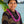 A man wearing a brown and black patterned shirt and an intricately folded round purple orchid lei, standing with Diamond Head, a palm tree, and the ocean in the background.