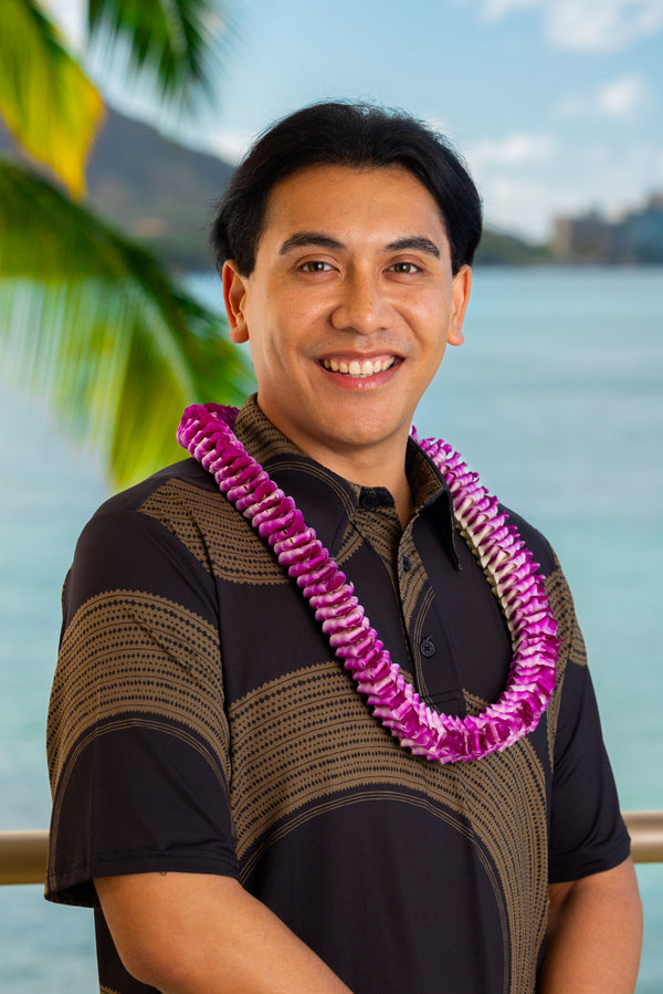 A man wearing a brown and black patterned shirt and an intricately folded round purple orchid lei, standing with Diamond Head, a palm tree, and the ocean in the background.