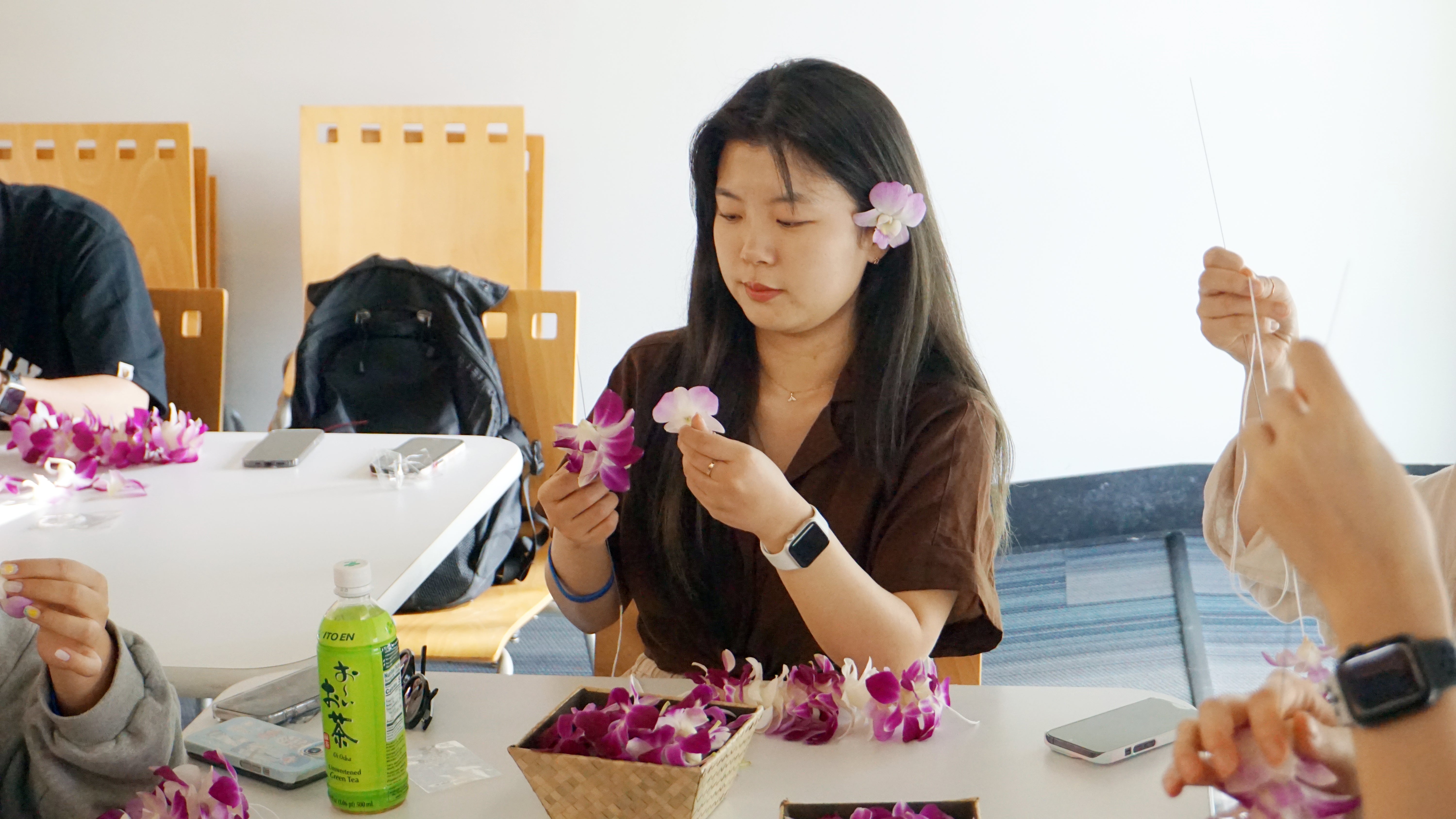 A girl at a table with a pink orchid in her hair, sewing orchid blossoms into a lei.