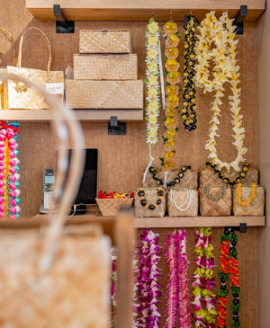 Inside a lei shop, pandan boxes stacked with orchid leis, silk plumeria leis, and kukui nut leis hanging above, and shell leis displayed in small lauhala boxes on shelves.