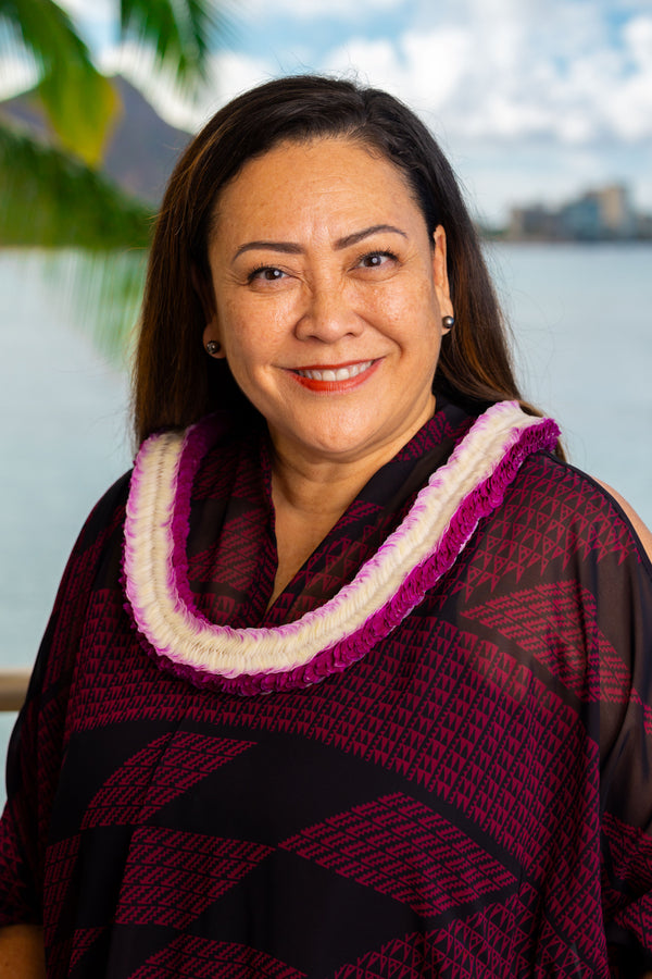 A woman wearing a black and deep magenta patterned shirt with a flat purple and white orchid lei, standing in front of Diamond Head, a palm tree, and the ocean.
