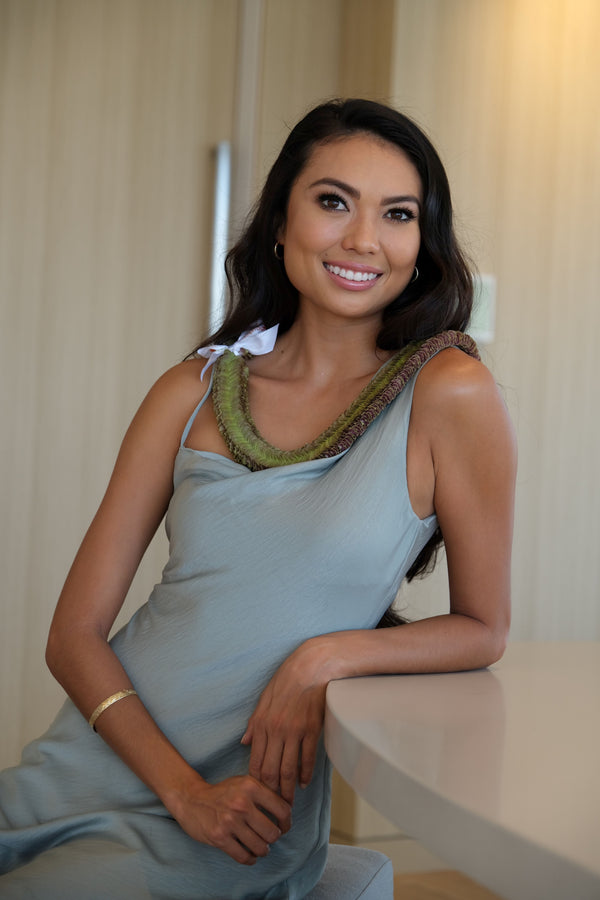 A seated woman in a teal dress wearing a jade green orchid lei with a white bow, resting her elbow on a table.