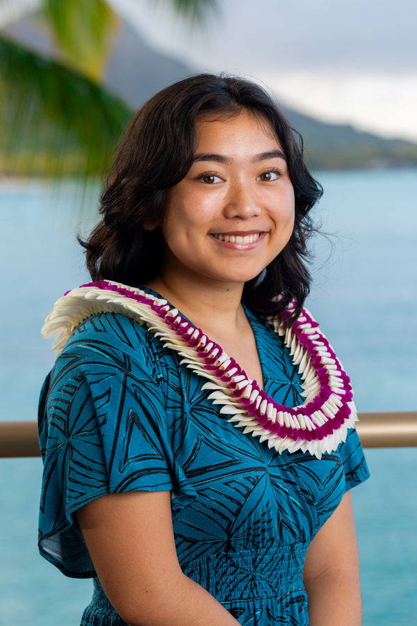 A young girl wearing a deep turquoise and black patterned dress and a flat orchid lei made of white and deep purple petals, standing with Diamond Head, a palm tree, and the ocean in the background.