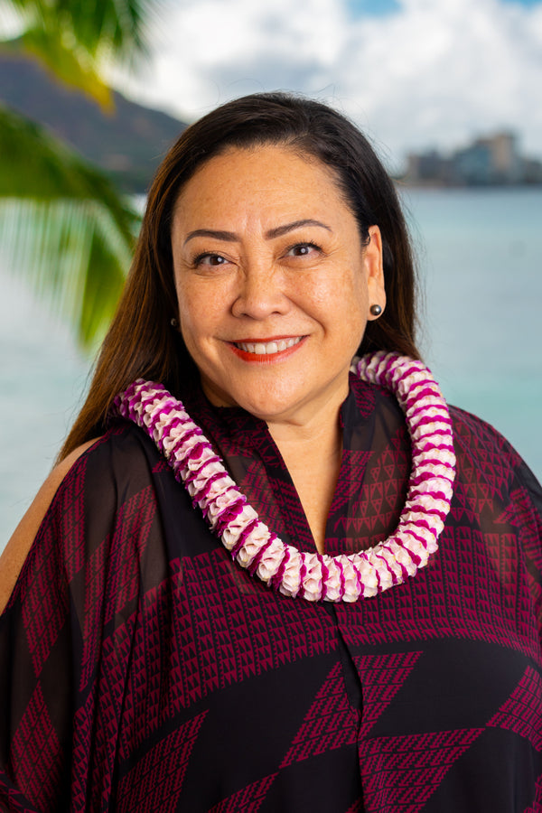 Woman wearing a deep magenta and black patterned shirt and a two-shaded purple spiral orchid lei, with Diamond Head, a palm tree, and the ocean in the background.
