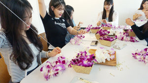 Korean girls sewing orchid leis, holding long strings with orchid blossoms in three different colors placed in baskets on the table in front of them.