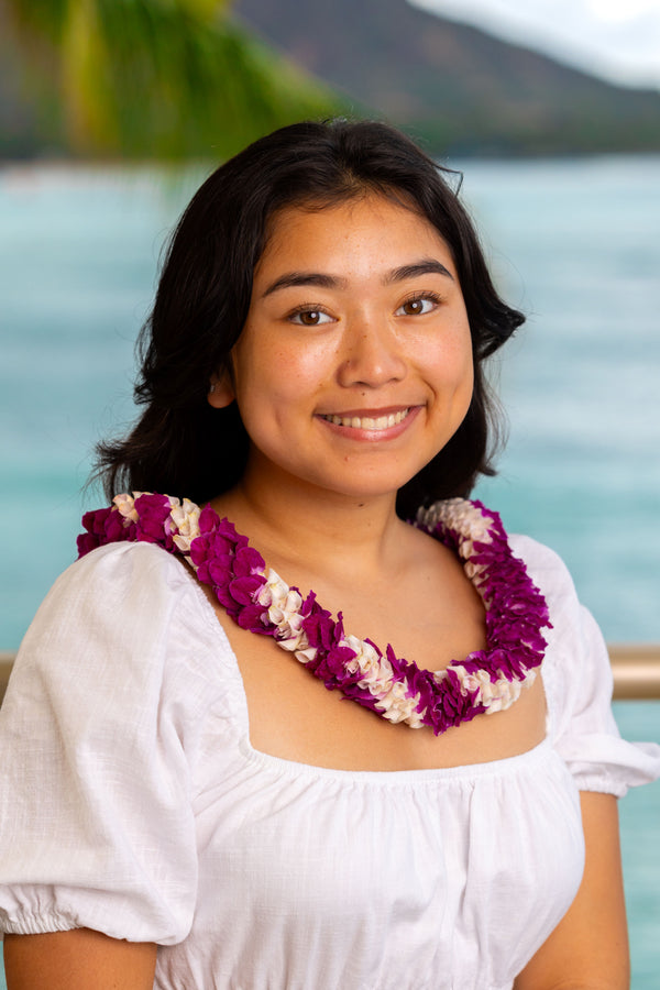 Young girl wearing a white dress and a two-colored purple spiral orchid lei, with Diamond Head, a palm tree, and the ocean in the background.