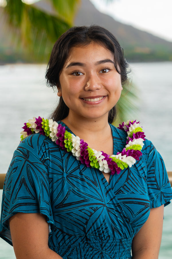 Young girl wearing a dark turquoise and black patterned dress and a spiral orchid lei in purple, white, and green, with Diamond Head, a palm tree, and the ocean in the background.