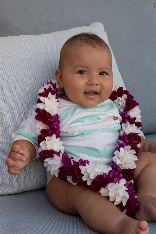 Baby leaning on a pillow, wearing a deep purple and white orchid lei.