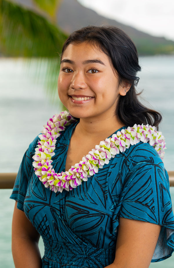 Teenage girl wearing a dark turquoise and black patterned dress and a spiral lei made of fresh orchid petals in purple, white, and green, with Diamond Head, a palm tree, and the ocean in the background.
