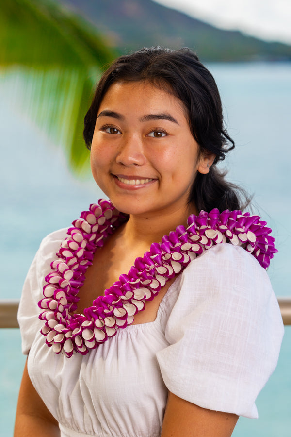 Young girl wearing a white dress and a purple and white orchid lei with white petals overlapping the purple, standing with Diamond Head, a palm tree, and the ocean in the background.