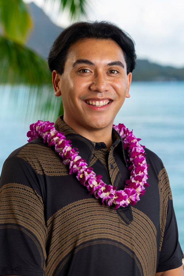 Man wearing a black and brown patterned shirt and a unique deluxe round purple orchid lei, with Diamond Head, a palm tree, and the ocean in the background.