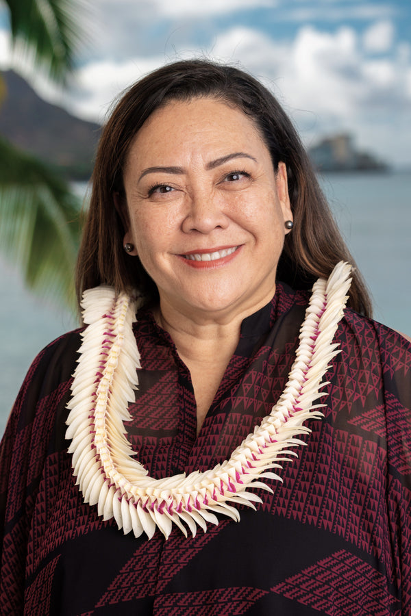 A woman wearing a deep magenta and black patterned shirt and a white orchid lei with subtle hints of deep purple, standing in front of Diamond Head, a palm tree, and the ocean in the background.