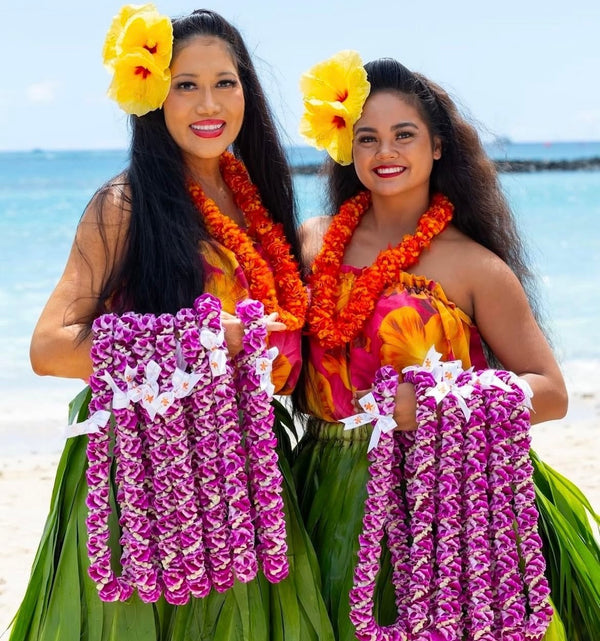 Two woman hula dancers with yellow hibiscus flowers in their hair, wearing orange leis and carrying numerous purple orchid leis adorned with white bows, with the ocean visible in the background.