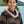 A man wearing a black and brown patterned shirt and a premium spiral orchid lei in white and purple, standing outdoors with Diamond Head, a palm tree, and the ocean in the background.