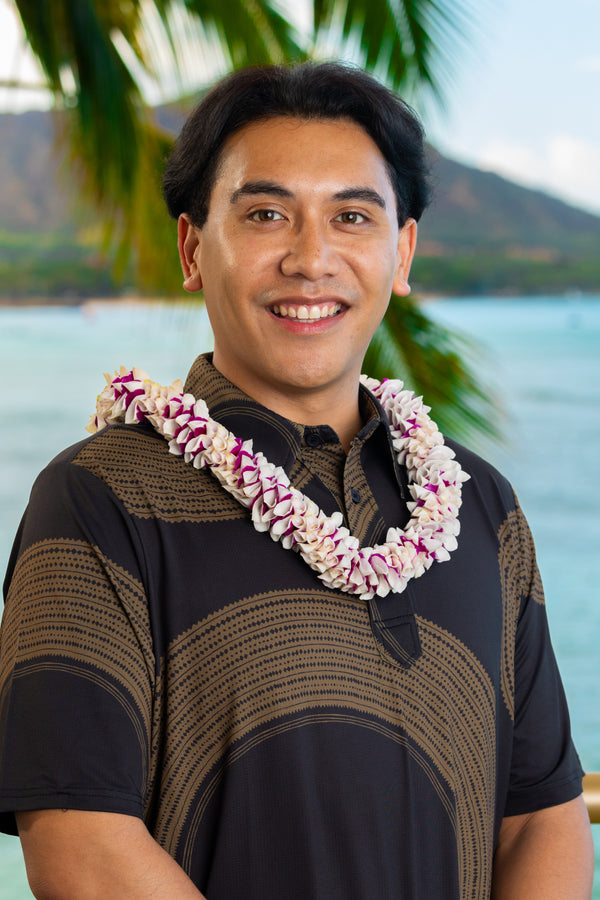 A man wearing a black and brown patterned shirt and a premium spiral orchid lei in white and purple, standing outdoors with Diamond Head, a palm tree, and the ocean in the background.