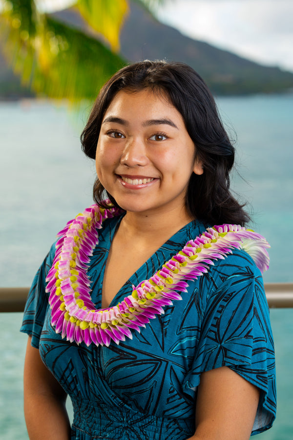 A girl wearing a deep turquoise and black patterned dress and a deluxe flat lei made of folded and flared orchid petals in purple and green, standing outdoors with Diamond Head, a palm tree, and the ocean in the background.