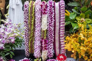 Display table featuring a sakura dendrobium arrangement on the left, a yellow mokara arrangement on the right, and seven premium orchid leis of various styles and colors arranged in the center.