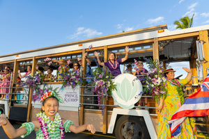 A trolley passing decorated with bunches of flowers, with people waving Hawaiian flags and wearing purple orchid leis.